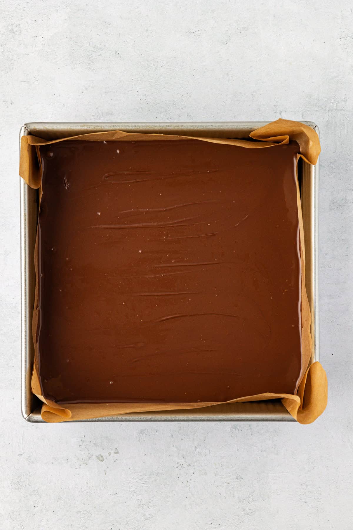 over head view of a square pan lined with parchment paper with a smooth chocolate layer in the pan