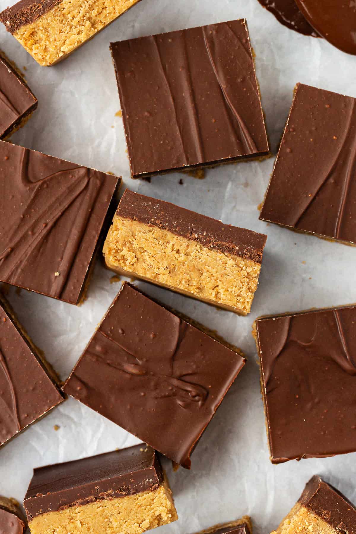 peanut butter bars scattered all around on white parchment paper, some laying flat and some on their sides showing the layers of peanut butter and chocolate