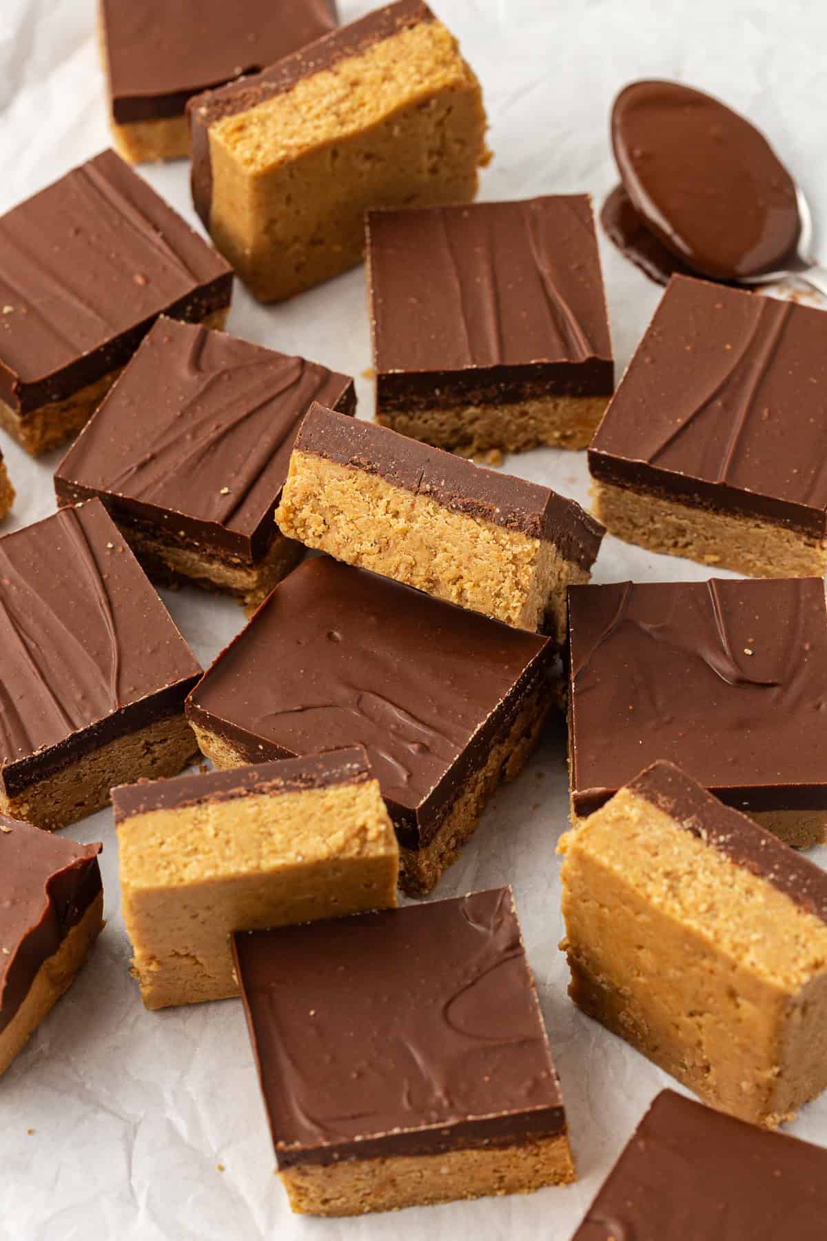 peanut butter bars scattered around on white parchment paper, some on their sides, some flat, some leaning on others, and one spoon of melted chocolate beside them