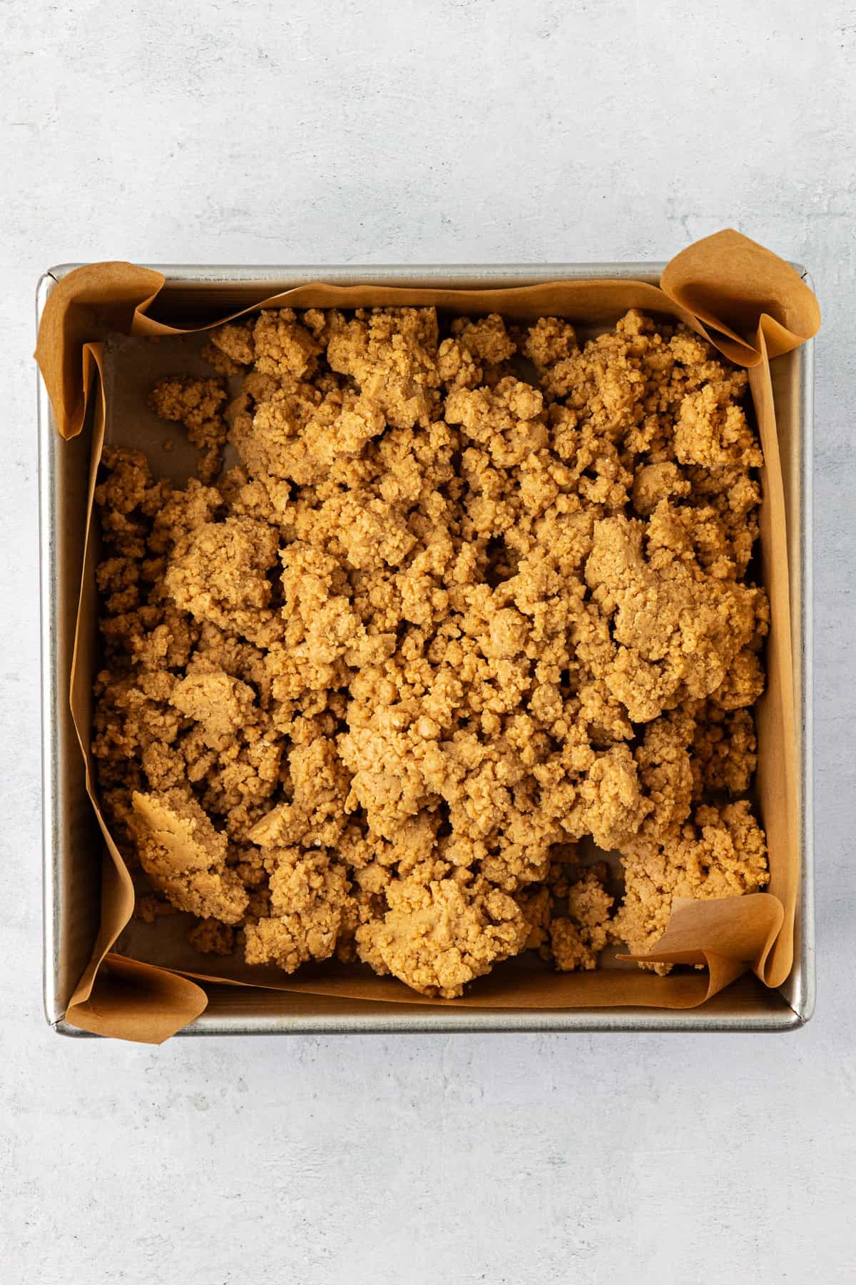 a square pan lined with brown parchment paper with a peanut butter and graham cracker mixture inside of it