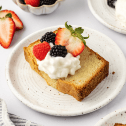 a slice of pound cake on a white plate topped with whipped cream and fresh berries, surrounding by more plates with slices, a bowl of berries and sliced strawberries