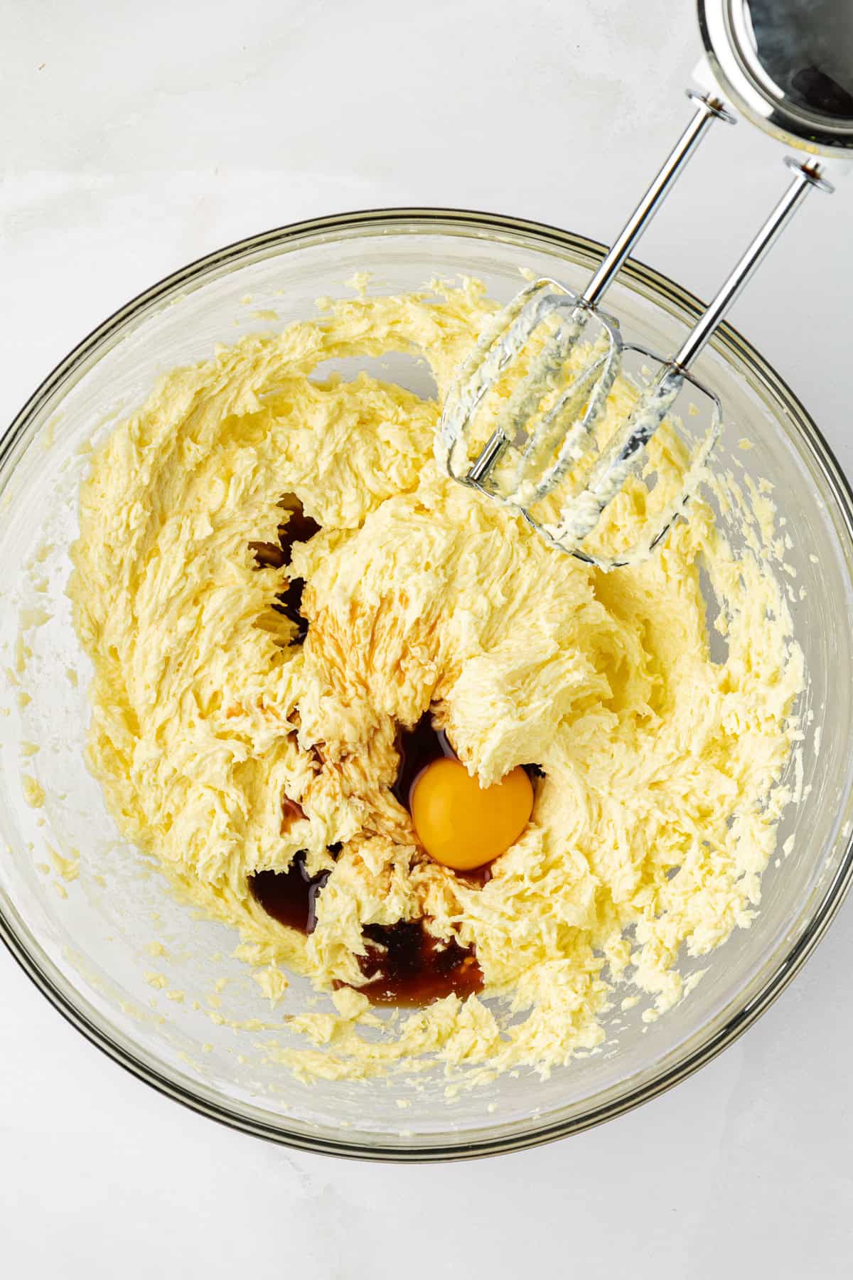 a mixture of butter and sugar with eggs and vanilla extract on top, not mixed in yet, with an electric mixer leaning on the side of the bowl
