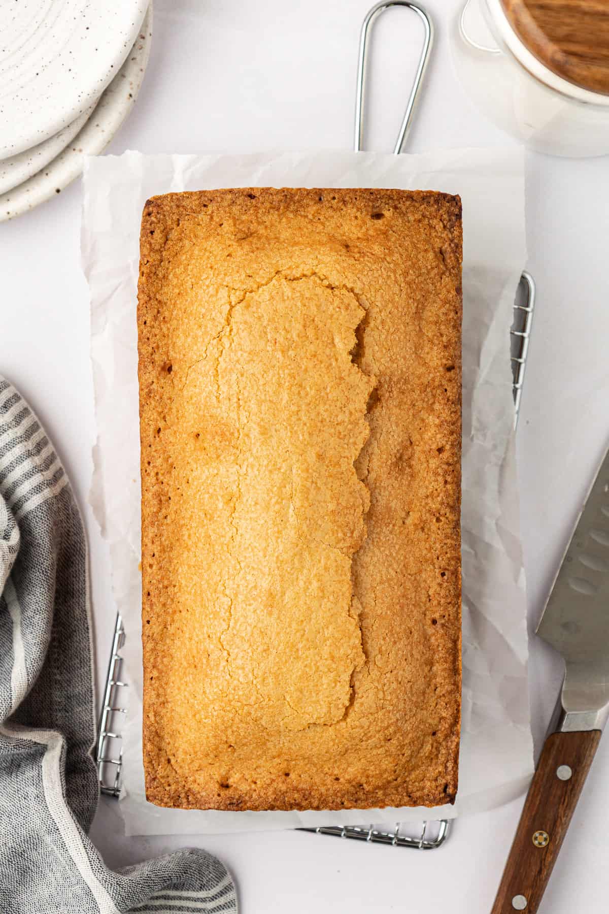 a whole pound cake sitting on parchment paper on a wire rack with a grey kitchen towel and a stack of plates to the left of it, a knife to the right