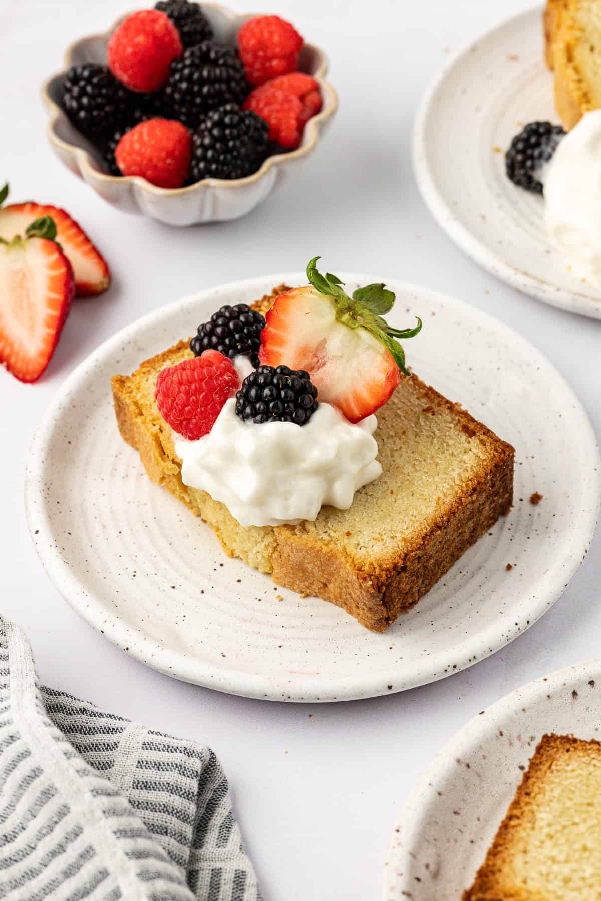 a slice of pound cake on a white plate topped with whipped cream and fresh berries, surrounding by more plates with slices, a bowl of berries and sliced strawberries