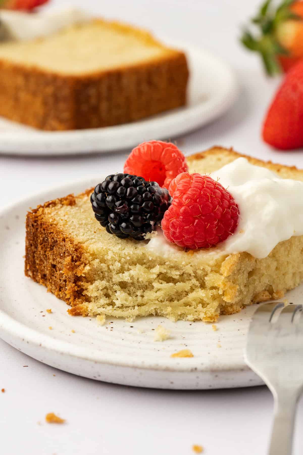 a slice of pound cake topped with whipped cream and fresh berries, with one bite missing a fork leaning on the plate, another slice on a plate and fresh strawberries in the background