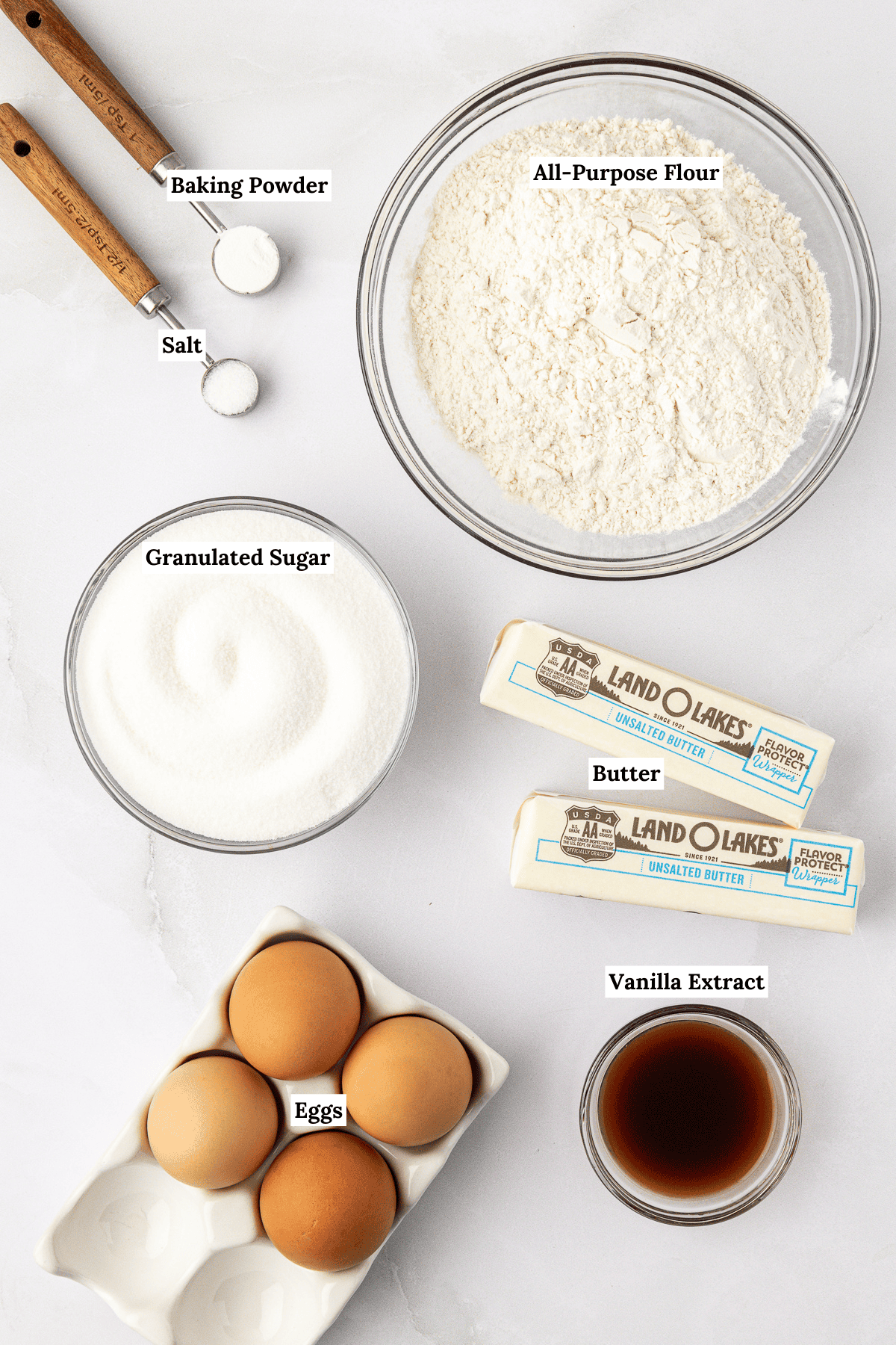 over head view of the ingredients for pound cake including baking powder, salt, all-purpose flour, two sticks of butter, granulated sugar, vanilla extract, and eggs
