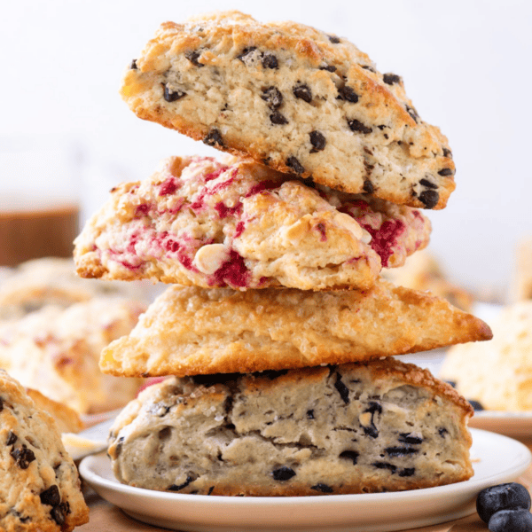Three scones stacked on a plate, each featuring different mix-ins: chocolate chips, raspberries, and blueberries. Other delicious scones are blurred in the background.