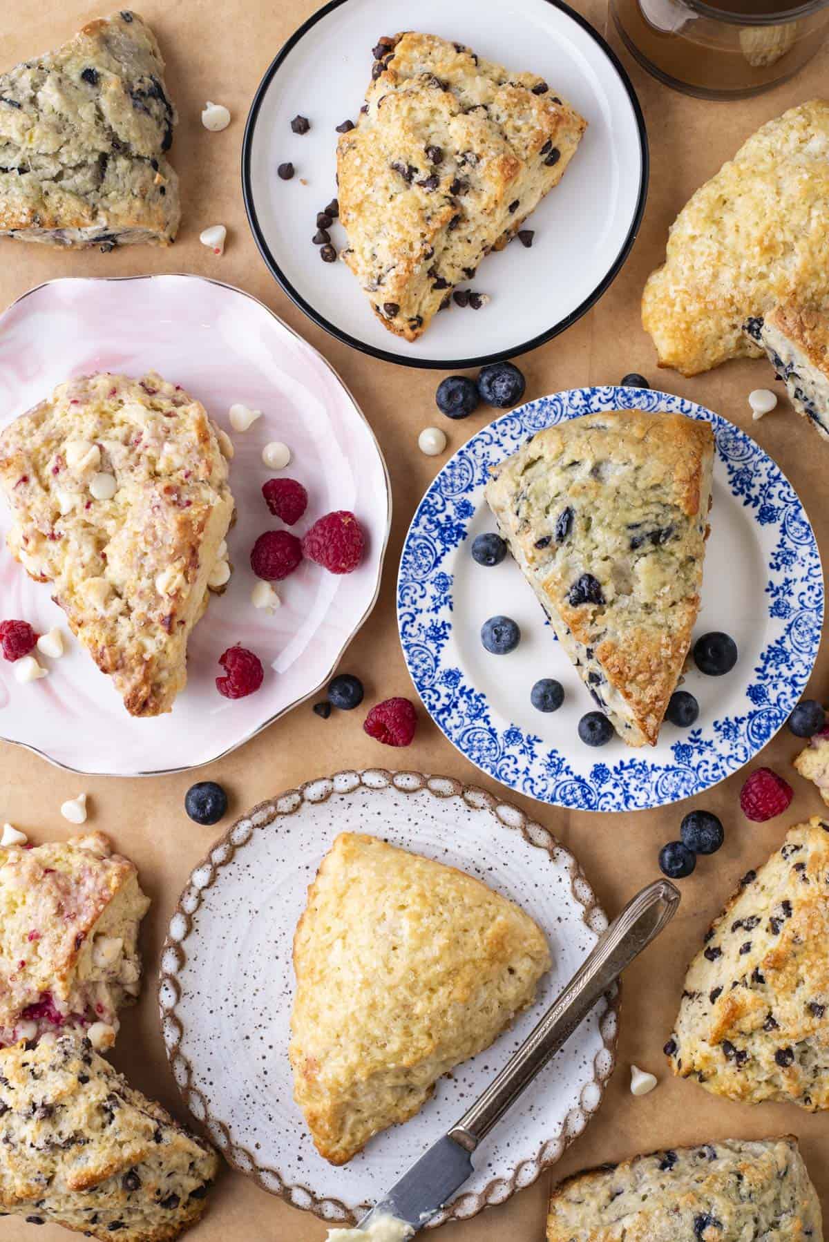 an arrangement of different flavored scones on colored plates including a raspberry scone on a pink plate, a blueberry scone on a blue plate, a chocolate chip scone on a white and black plate and a plain scone on a speckled plate, with fresh blueberries and raspberries and mini white chocolate chips scattered around