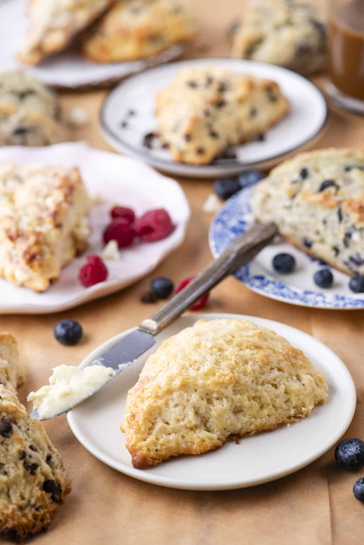 an assortment of scone flavors on plates with fresh berries sprinkled around and a butter knife with a bit of butter on it on the front plate with a plain scone