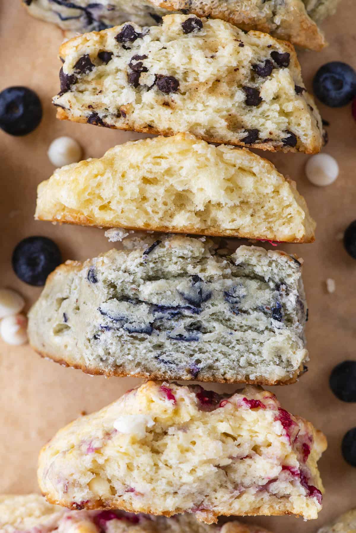 an assortment of scone flavors on their sides cut open showing the insides including chocolate chip, plain, blueberry and white chocolate raspberry, with fresh berries and white chocolate chips sprinkled around