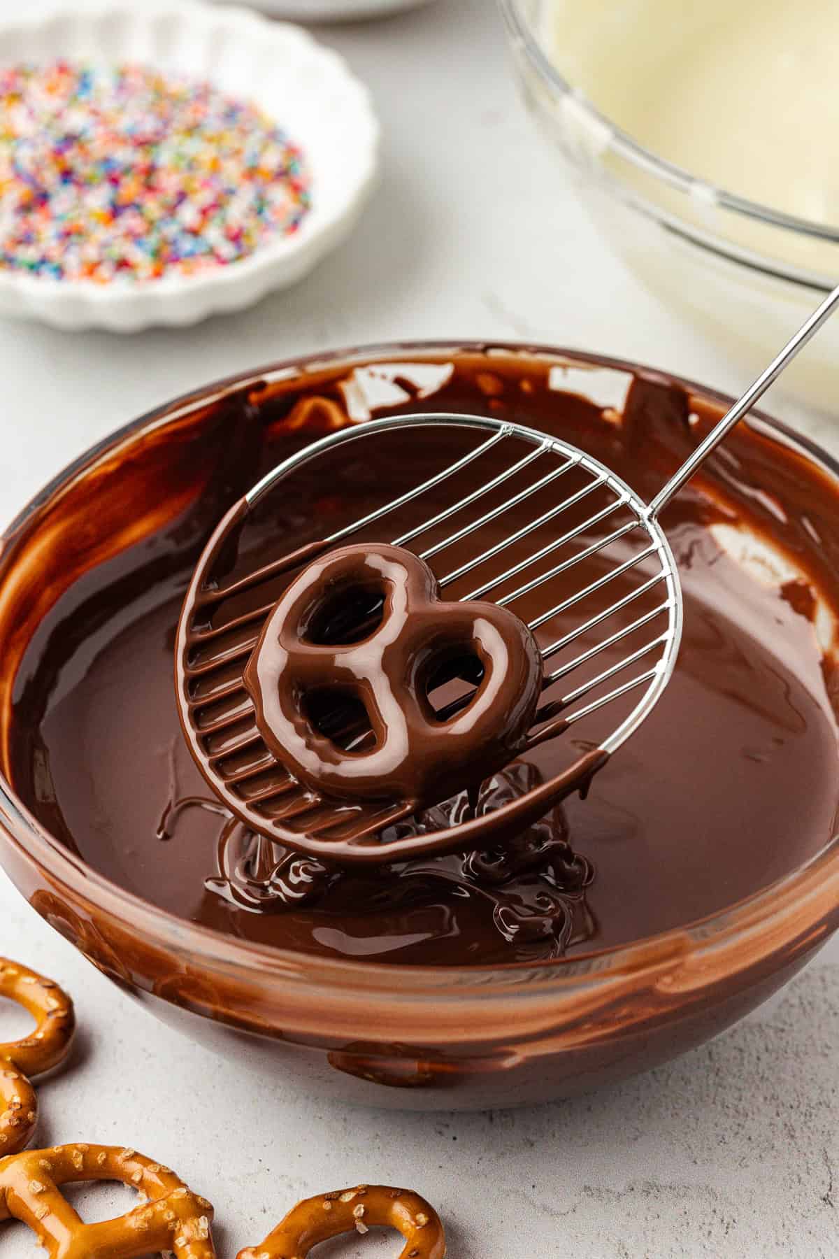 a chocolate covered pretzel being lifted out of a clear glass bowl full of melted chocolate, with a few pretzels scattered around, a white bowl of rainbow sprinkles and a clear glass bowl of melted white chocolate in the background