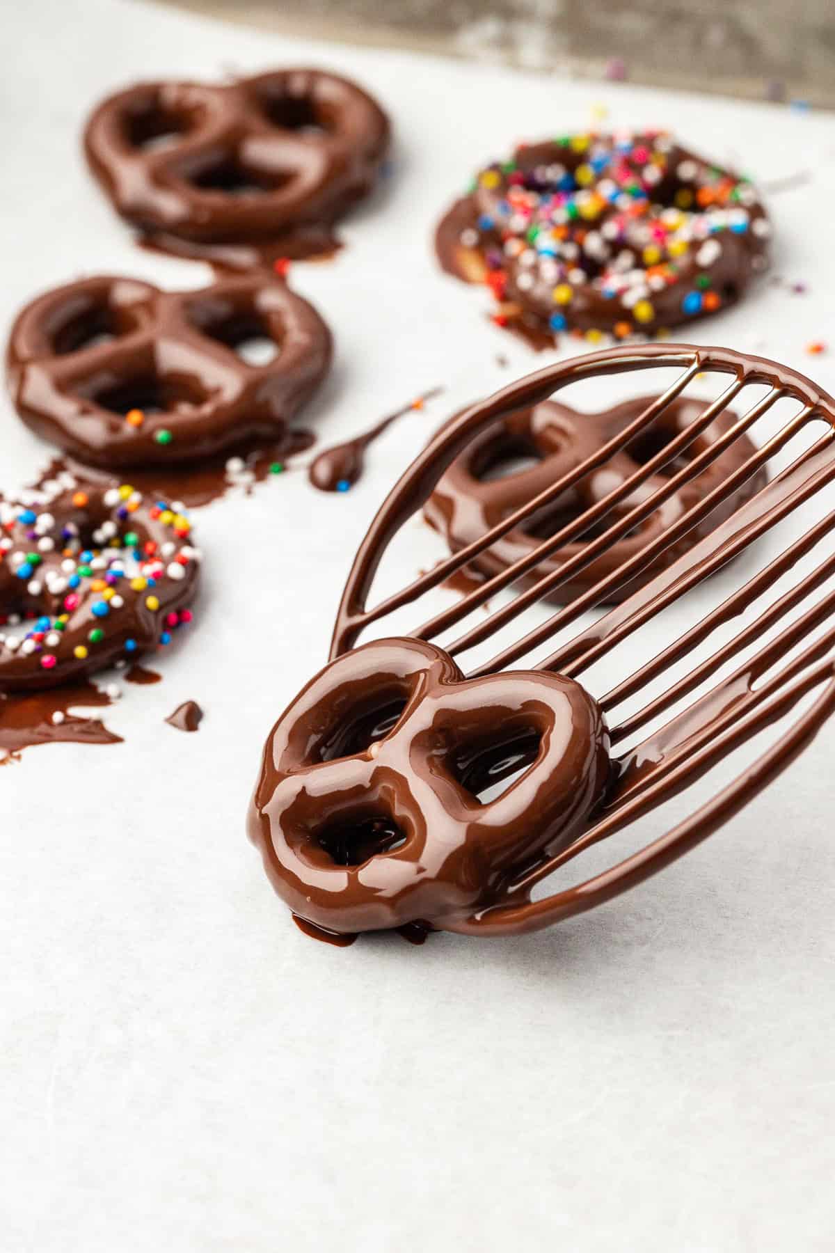 a chocolate coated pretzel being placed on a baking sheet beside other chocolate covered pretzels, two of which also have rainbow sprinkles on them