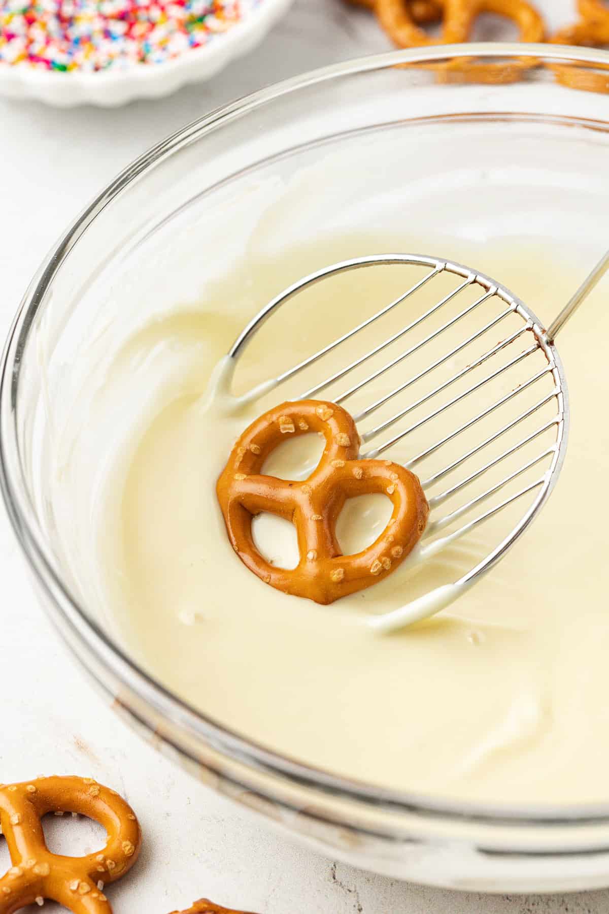 a twist pretzel being dipped into a clear glass bowl of white chocolate with more pretzels scattered around and a bowl of rainbow sprinkles in the background