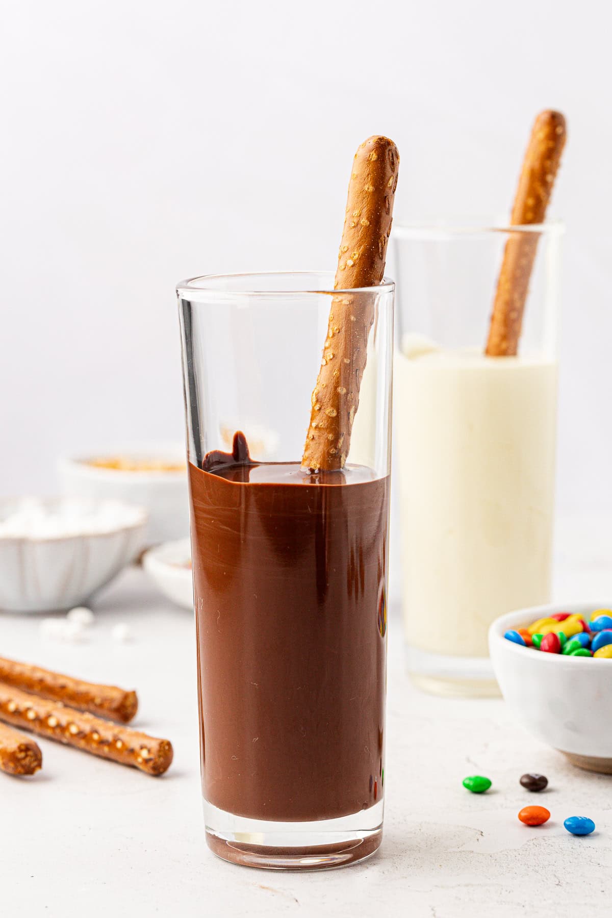 two tall glasses, one filled with melted chocolate and the other filled with melted white chocolate, each with a pretzel rod stuck into them, a bowl of m&ms, and more pretzel rods and m&ms scattered around
