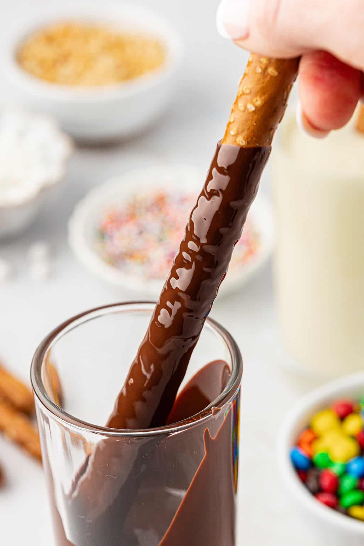 a pretzel rod being dipped into a glass of melted chocolate with several small bowls of different toppings like m&ms and sprinkles in the background