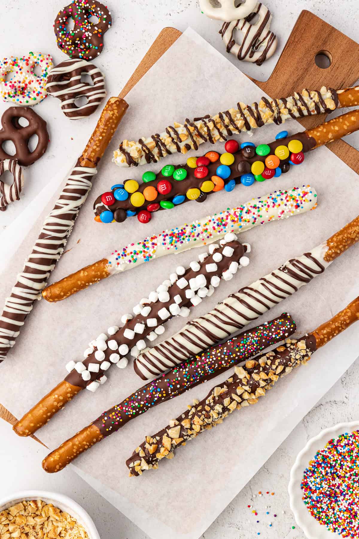 an assortment of chocolate covered pretzel rods and twist pretzels on a cutting board with white paper on it, with a bowl of crushed nuts and a bowl of sprinkles beside it