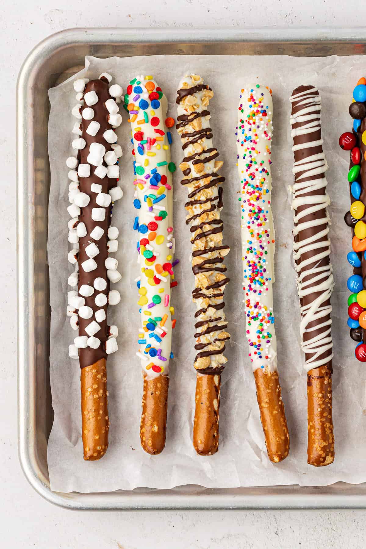 a row of chocolate covered pretzel rods on a baking sheet with different toppings: first chocolate with mini marshmallows, next white chocolate with rainbow sprinkles, next white chocolate with crushed nuts and chocolate drizzle, next white chocolate with mini rainbow sprinkles, next chocolate drizzled with white chocolate, and then chocolate with m&ms