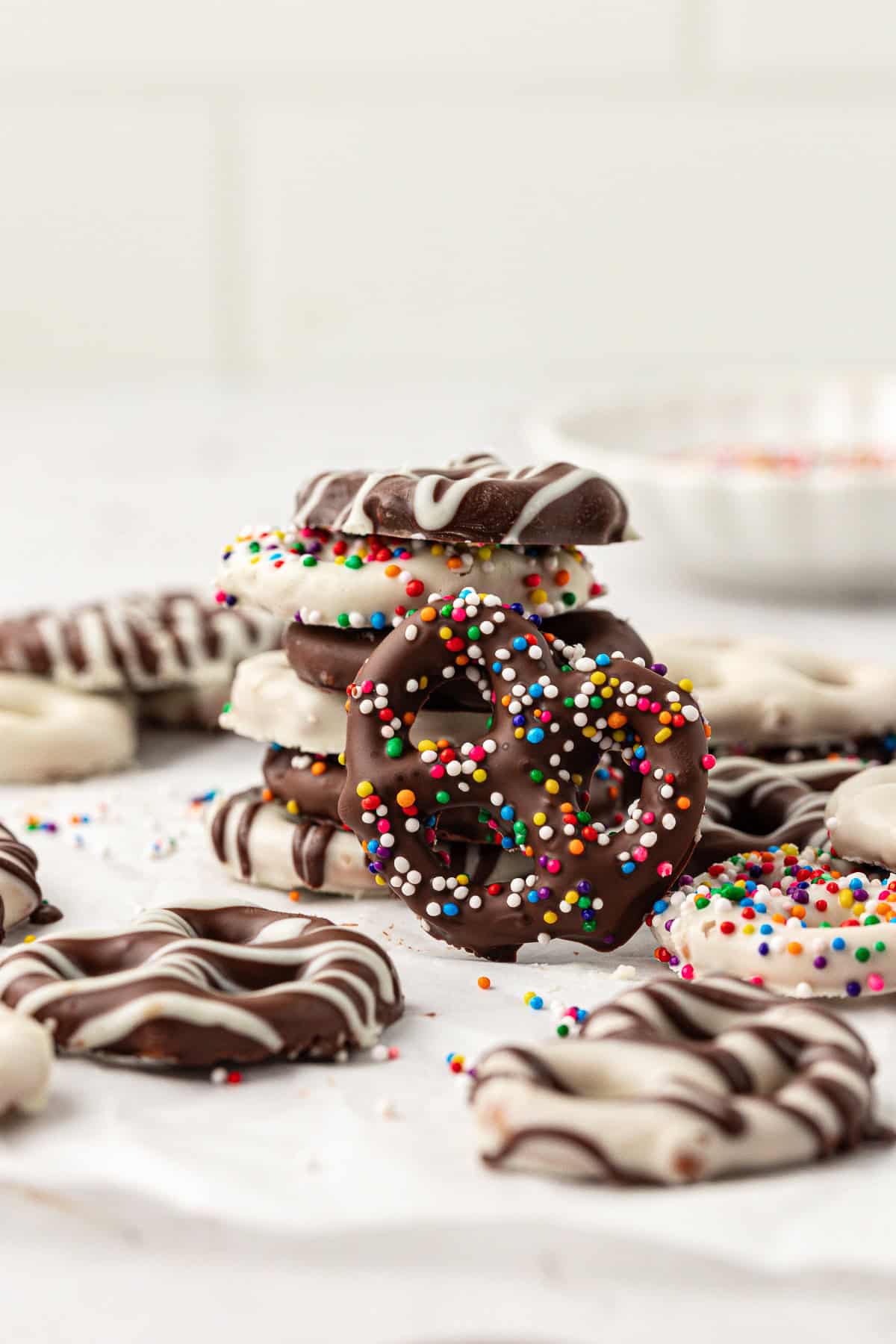 an assortment of chocolate covered pretzels stacked and scattered around including chocolate with white chocolate drizzle, white chocolate with chocolate drizzle, and both chocolate and white chocolate coated in rainbow sprinkles