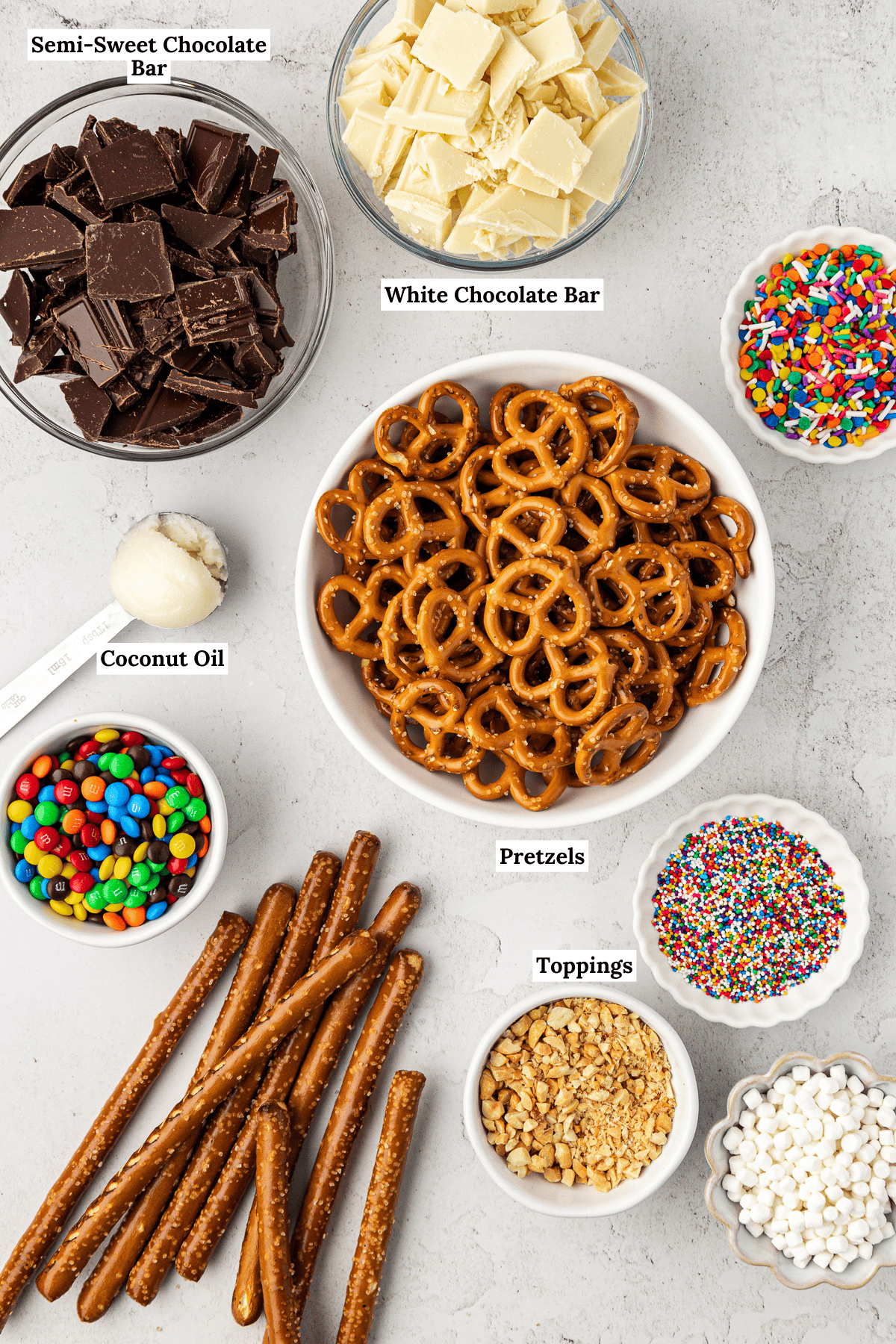 over head view of chocolate covered pretzels including semi-sweet chocolate bars, white chocolate bars, coconut oil, rainbow sprinkles, m&ms, crushed nuts and mini marshmallows, twist pretzels and pretzel rods