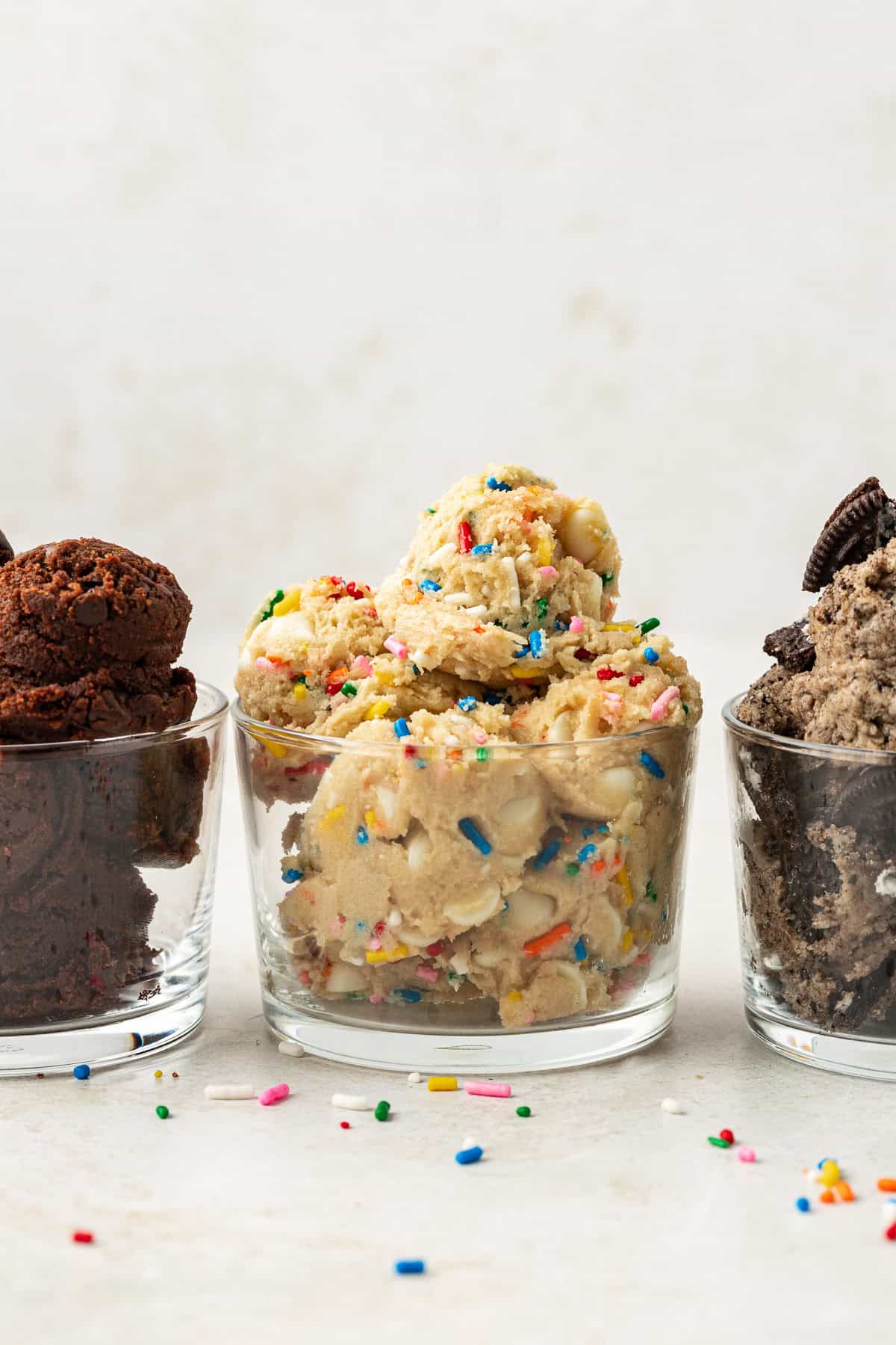 three round glass containers full of edible cookie dough with chocolate on the left, funfetti in the middle and oreo flavor on the right, with rainbow sprinkles scattered around