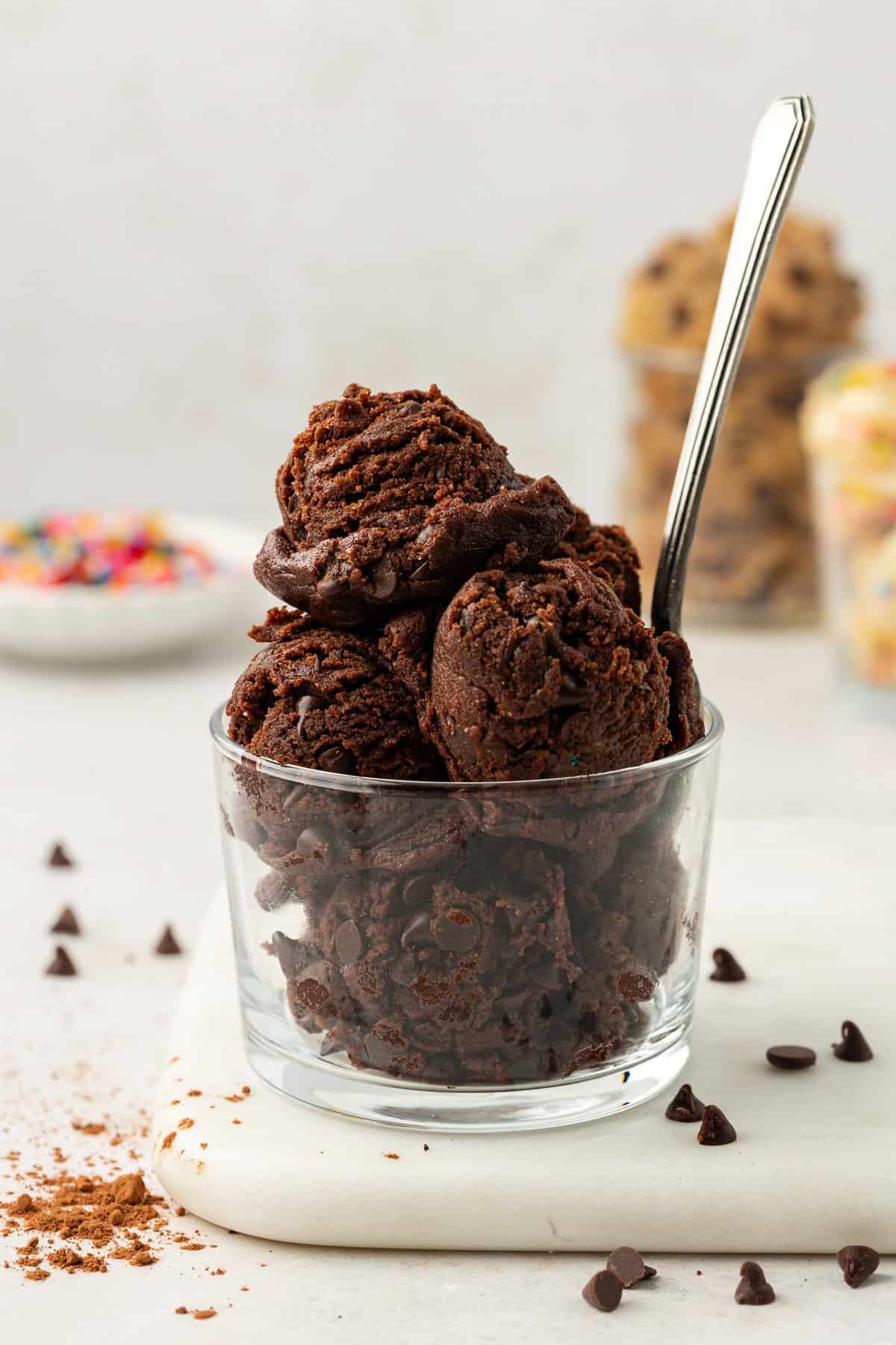 edible chocolate cookie dough balls in a clear glass container with a spoon on top of a white cutting board with chocolate chips sprinkled around and more cookie dough flavors in the background in clear glass containers