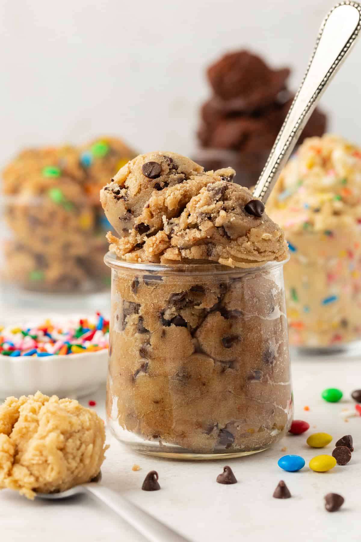 a clear glass jar filled with chocolate chip cookie dough with a spoon in it with m&ms and chocolate chips scattered around, more flavors of cookie dough in glass containers in the background and spoon full of peanut butter cookie dough in the front