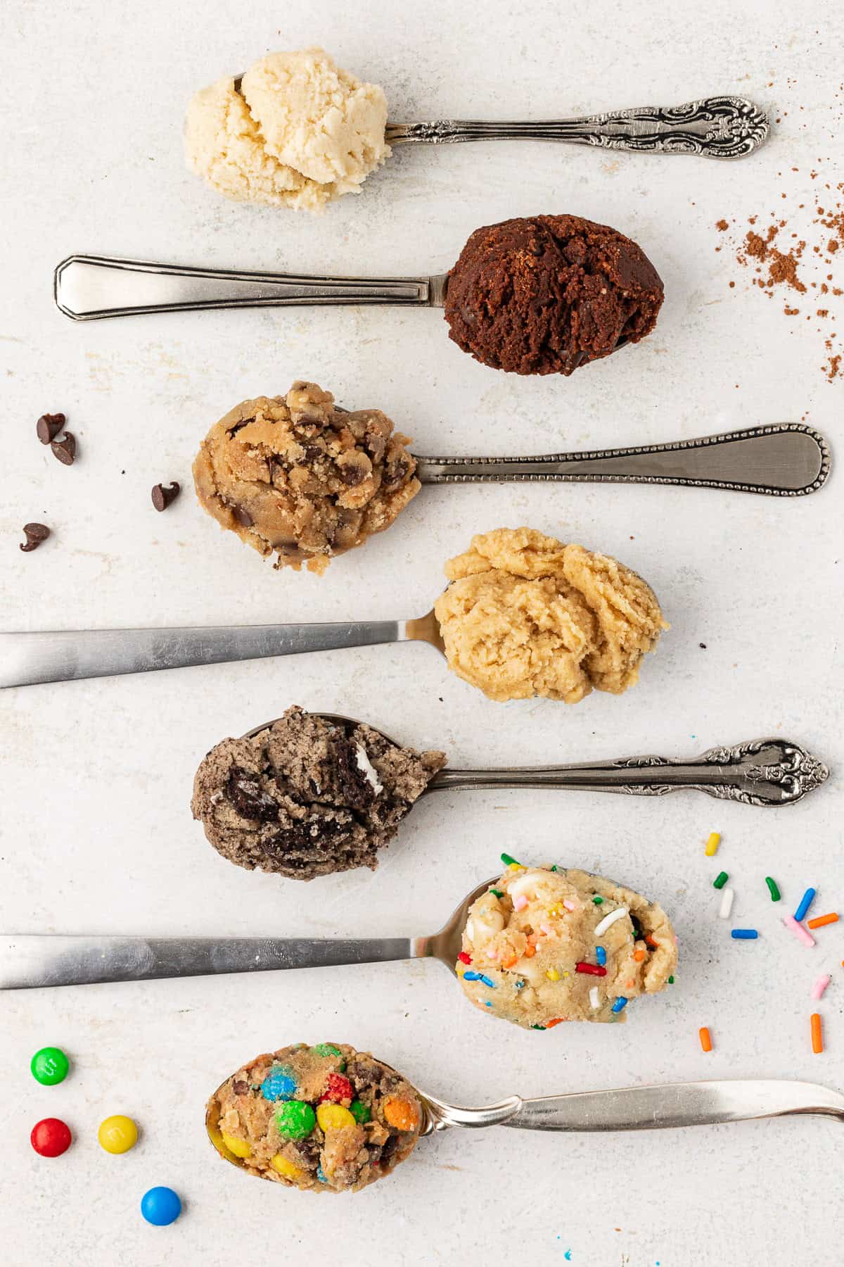 seven spoons alternating directions laying flat with edible cookie dough in them starting with sugar cookie, then chocolate, chocolate chip, peanut butter, oreo, funfettie and cookie monster at the bottom, with m&ms, sprinkles, chocolate chips, and cocoa powder sprinkled around