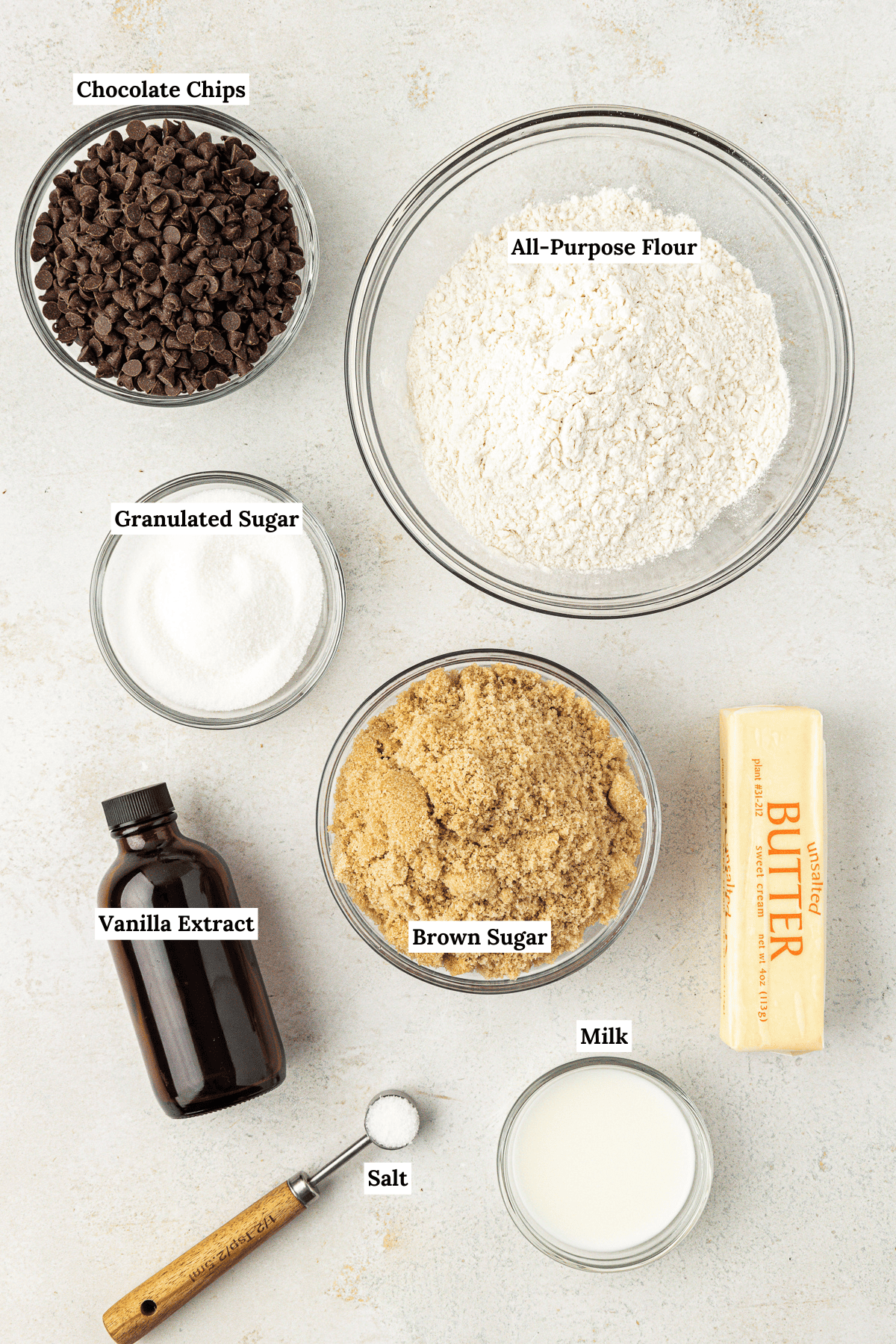 overhead view of edible cookie dough ingredients including chocolate chips, all-purpose flour, granulated sugar, brown sugar, vanilla extract, butter, milk and salt