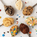 seven spoons in a circle with different flavors of edible cookie dough with m&ms, chocolate chips and sprinkles scattered around