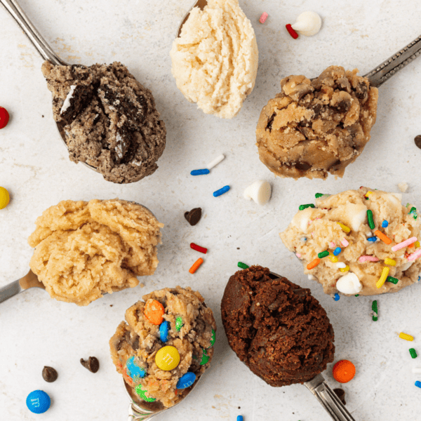seven spoons in a circle with different flavors of edible cookie dough with m&ms, chocolate chips and sprinkles scattered around