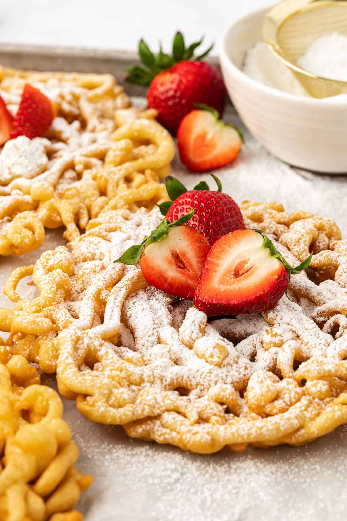close up of funnel cakes on a baking sheet topped with powdered sugar and fresh strawberries with a white bowl of powdered sugar and a sifter beside them