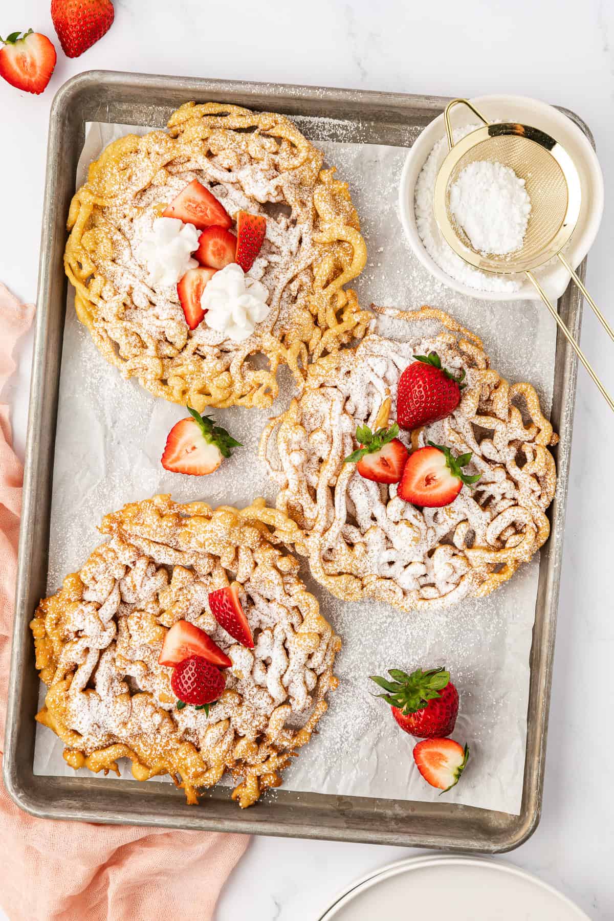 three funnel cakes on a baking sheet lined with parchment paper topped with powdered sugar and fresh strawberries with a pink cloth on the left, fresh strawberries scattered around and a small white bowl of powdered sugar with a sifter in it