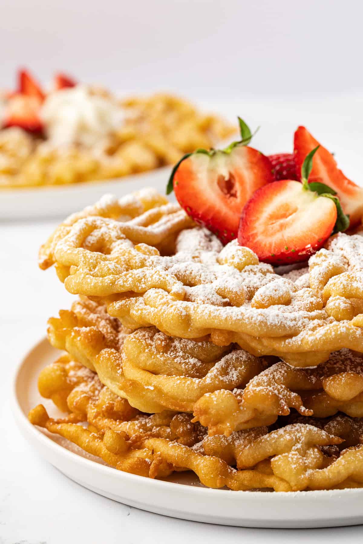 three layers of funnel cake on a white plate topped with powdered sugar and fresh strawberries with another funnel cake in the background