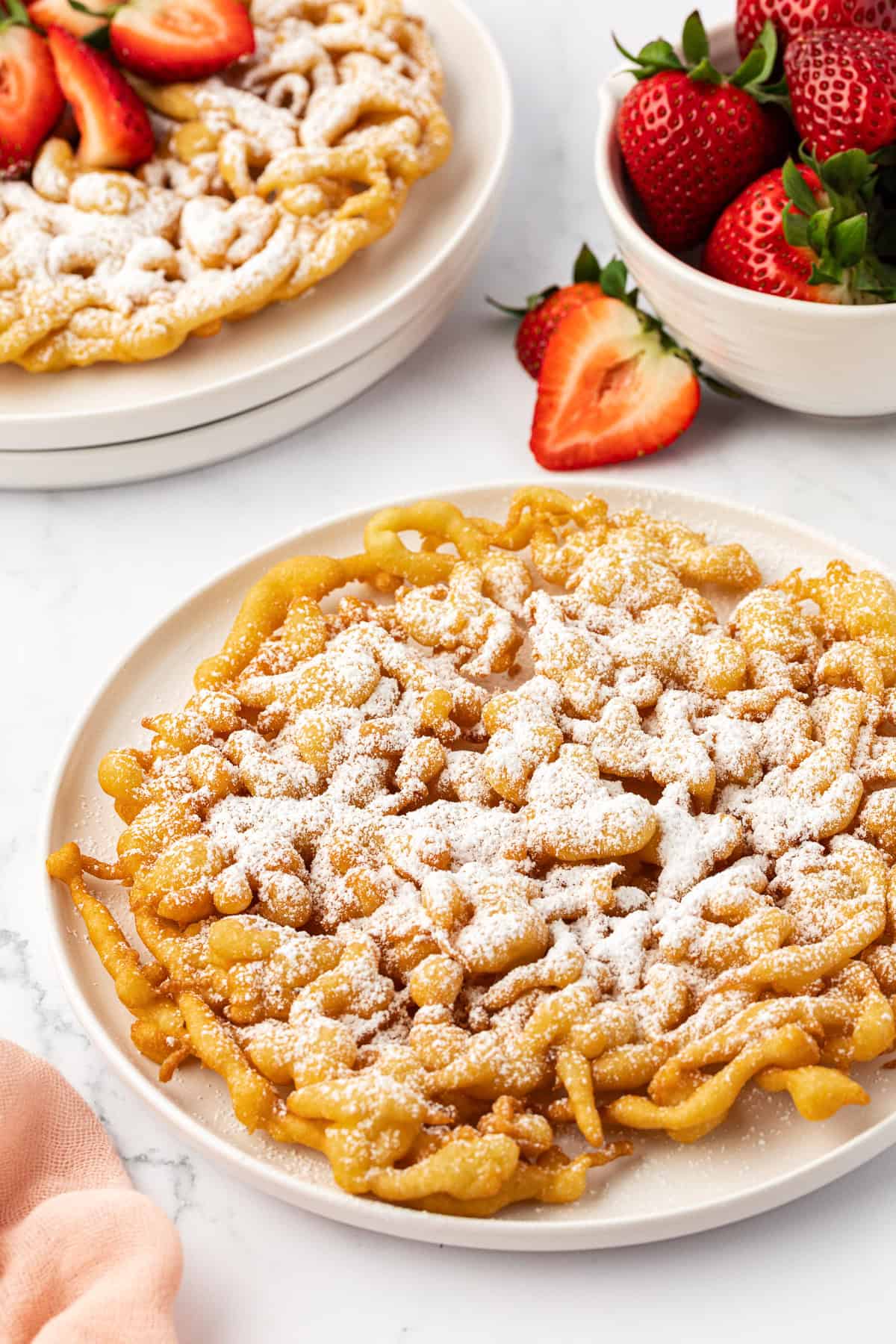two funnel cakes on white plates with the front one topped with powdered sugar and the back one topped with powdered sugar and fresh strawberries, and a bowl of whole strawberries on the right