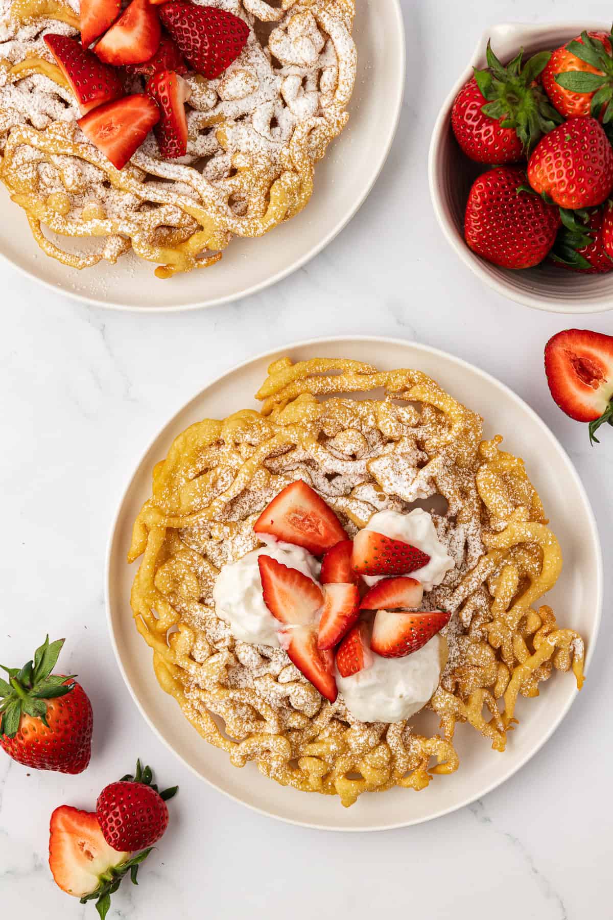 two funnel cakes on white plates topped with powdered sugar and fresh strawberries with fresh strawberries scattered around and a bowl of whole strawberries on the right
