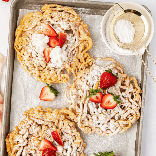 over head view of three funnel cakes on a baking sheet lined with parchment paper topped with powdered sugar and fresh strawberries and a bowl of powdered sugar with a sifter in it