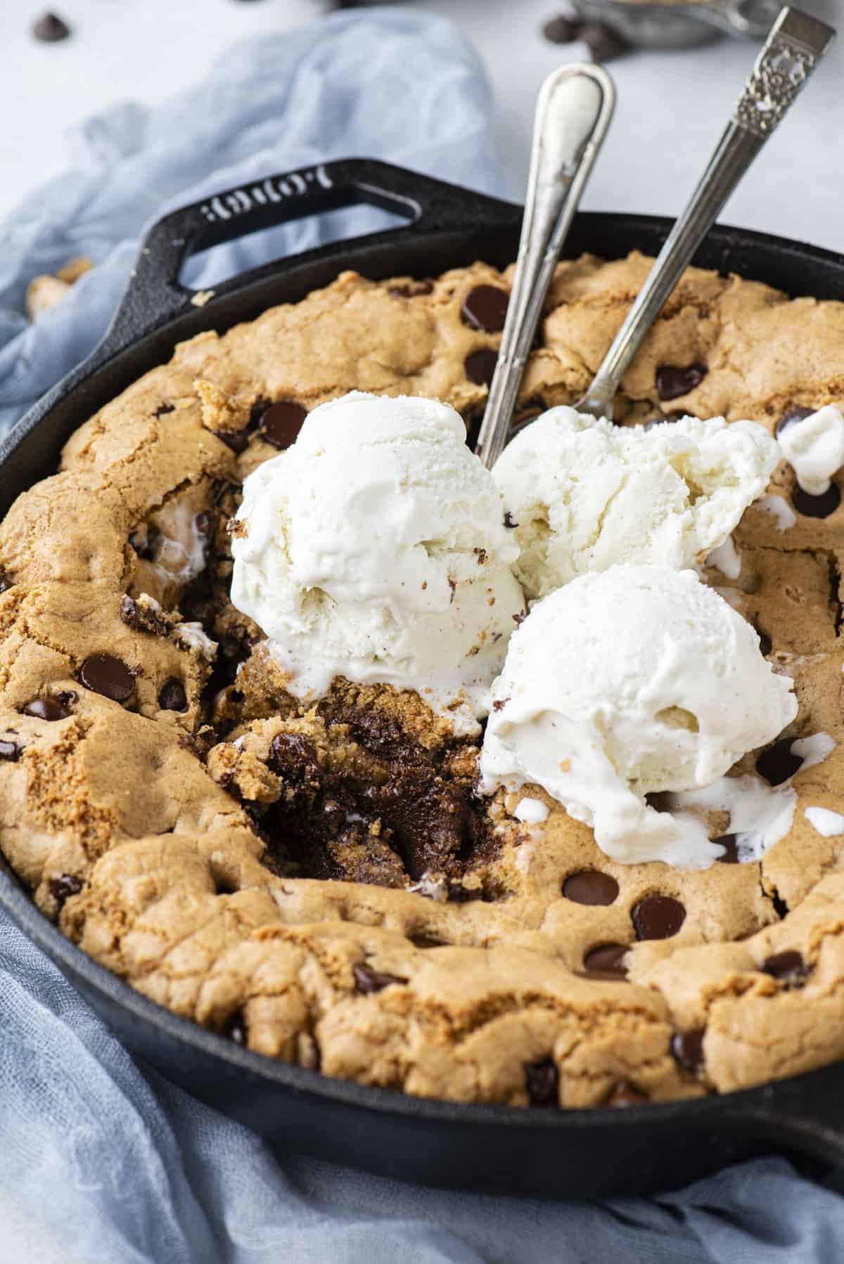 a cast iron skillet full of pizookie on top of a blue cloth, with three scoops of vanilla ice cream on top, bites missing out of the middle and two spoons stuck in the top of it