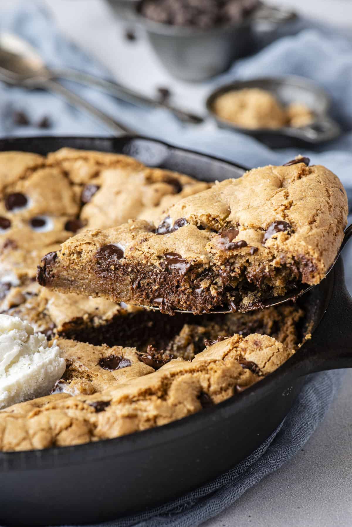 a slice of pizookie being lifted out of a cast iron skillet that is sitting on top of a blue cloth with chocolate chips and spoons in the background