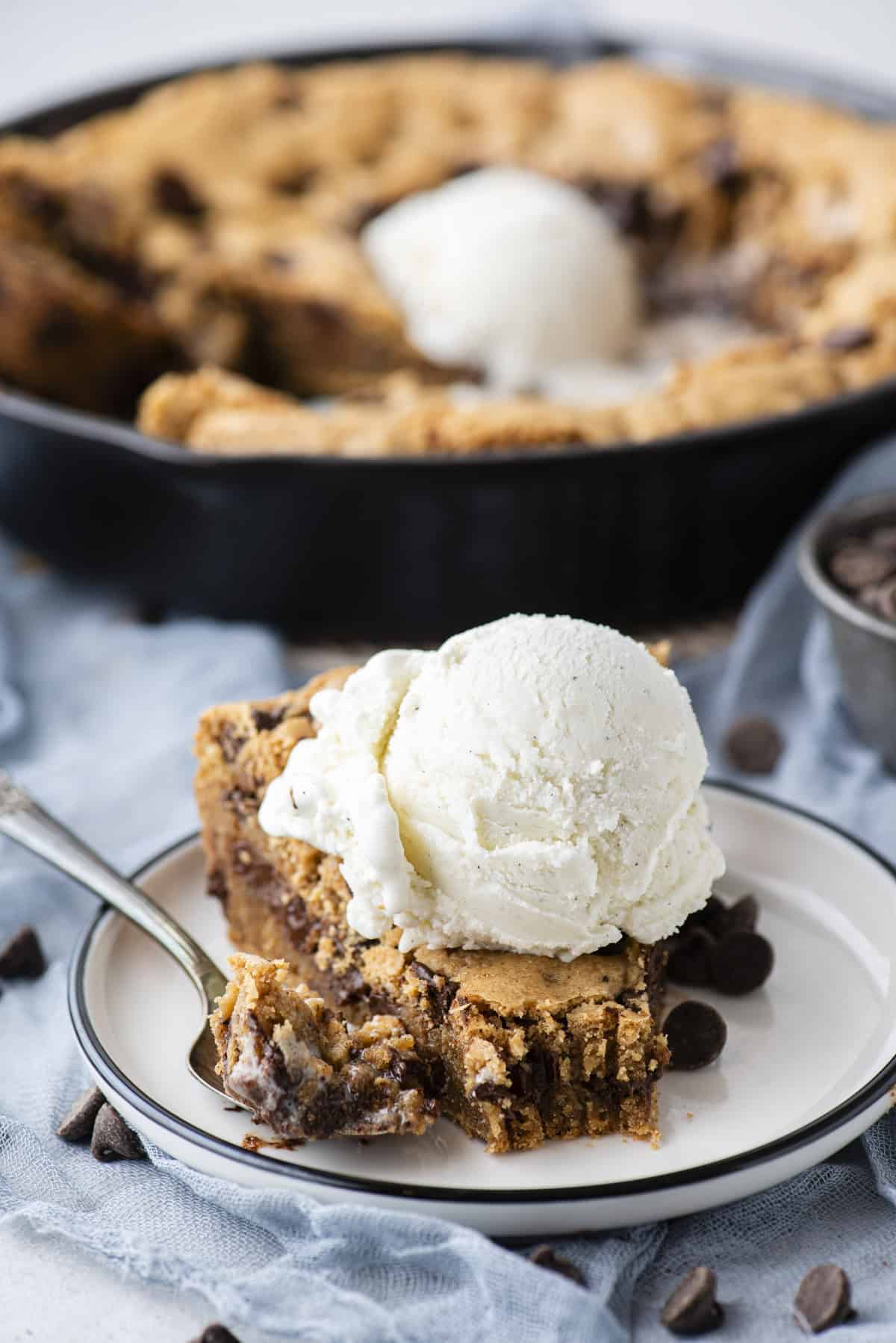 a slice of pizookie on a small white plate topped with a scoop of vanilla ice cream with a spoon beside the slice with a bite of pizookie on it, and a whole cast iron skillet of pizookie in the background, and chocolate chips scattered around