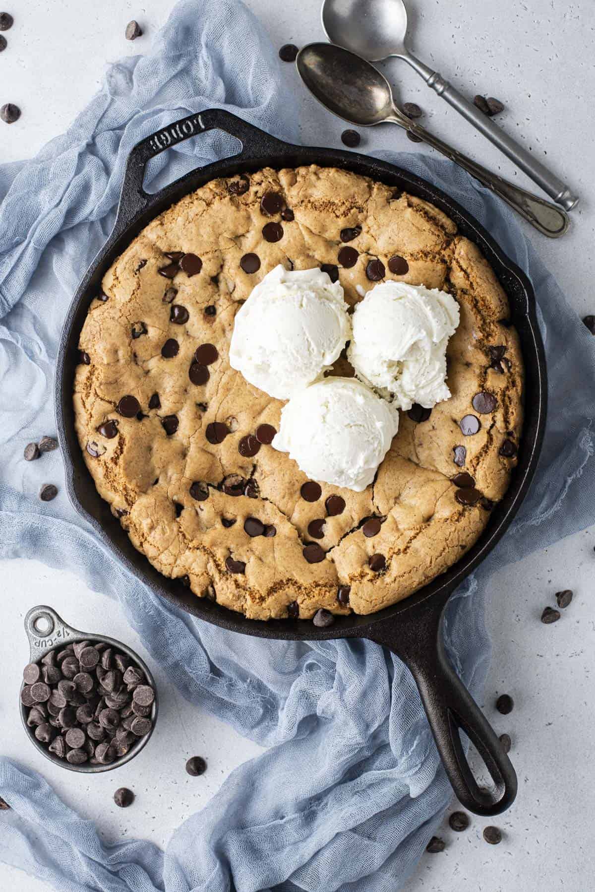 overhead view of a cast iron skillet full of pizookie with three scoops of vanilla ice cream on top, the skillet sitting on a blue cloth surrounded by chocolate chips scattered around, a cup of chocolate chips and two spoons