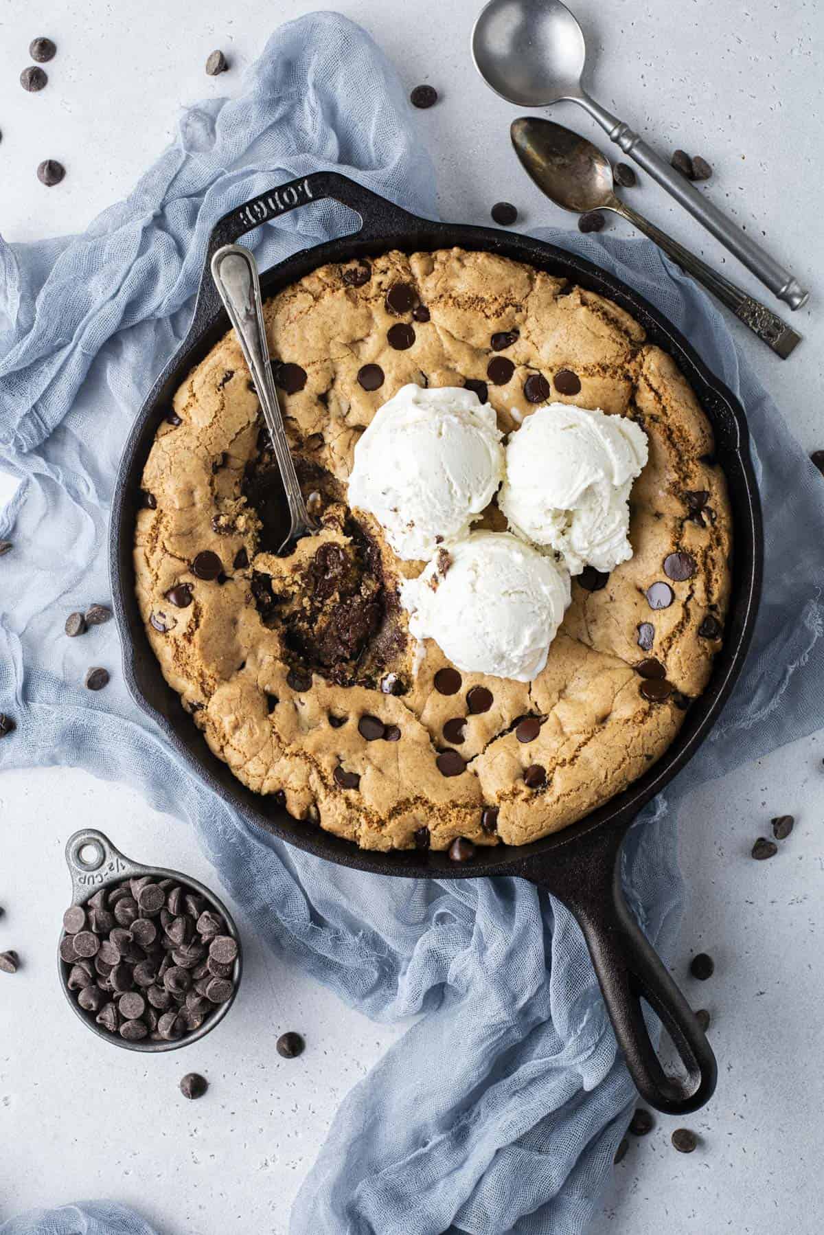 overhead view of a cast iron skillet full of a giant chocolate chip cookie (pizookie) and three scoop of vanilla ice cream, with a spoon in the middle and bites missing out, sitting on top of a blue cloth surrounded by a cup of chocolate chips, chocolate chips scattered around and two spoons