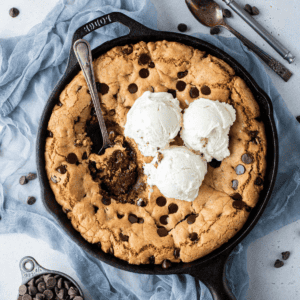 overhead view of a cast iron skillet full of a giant chocolate chip cookie (pizookie) and three scoop of vanilla ice cream, with a spoon in the middle and bites missing out, sitting on top of a blue cloth surrounded by a cup of chocolate chips, chocolate chips scattered around and two spoons