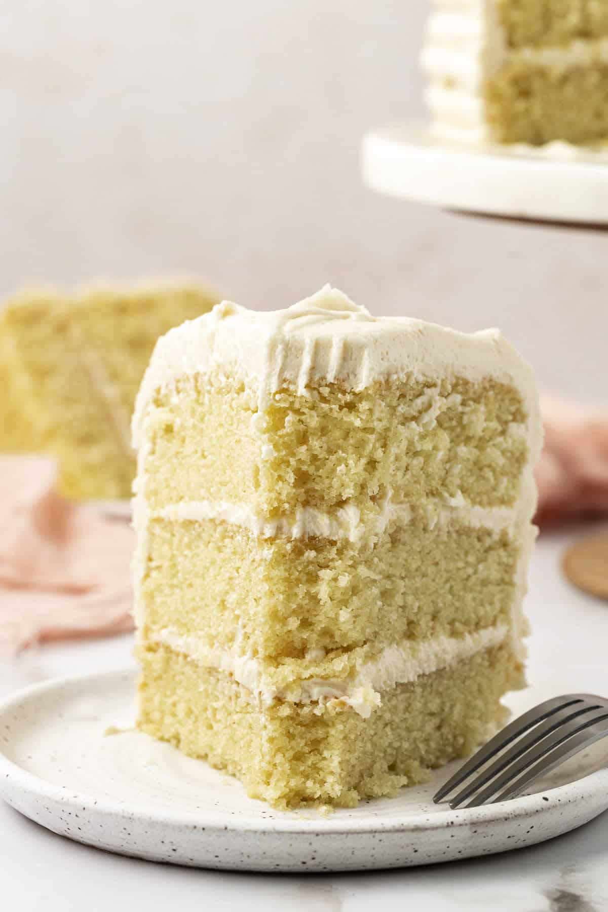 a slice of 3-layer vanilla cake on a white speckled plate with a fork leaning on the plate and a bite missing out of the cake slice and a cake stand with more cake in the background
