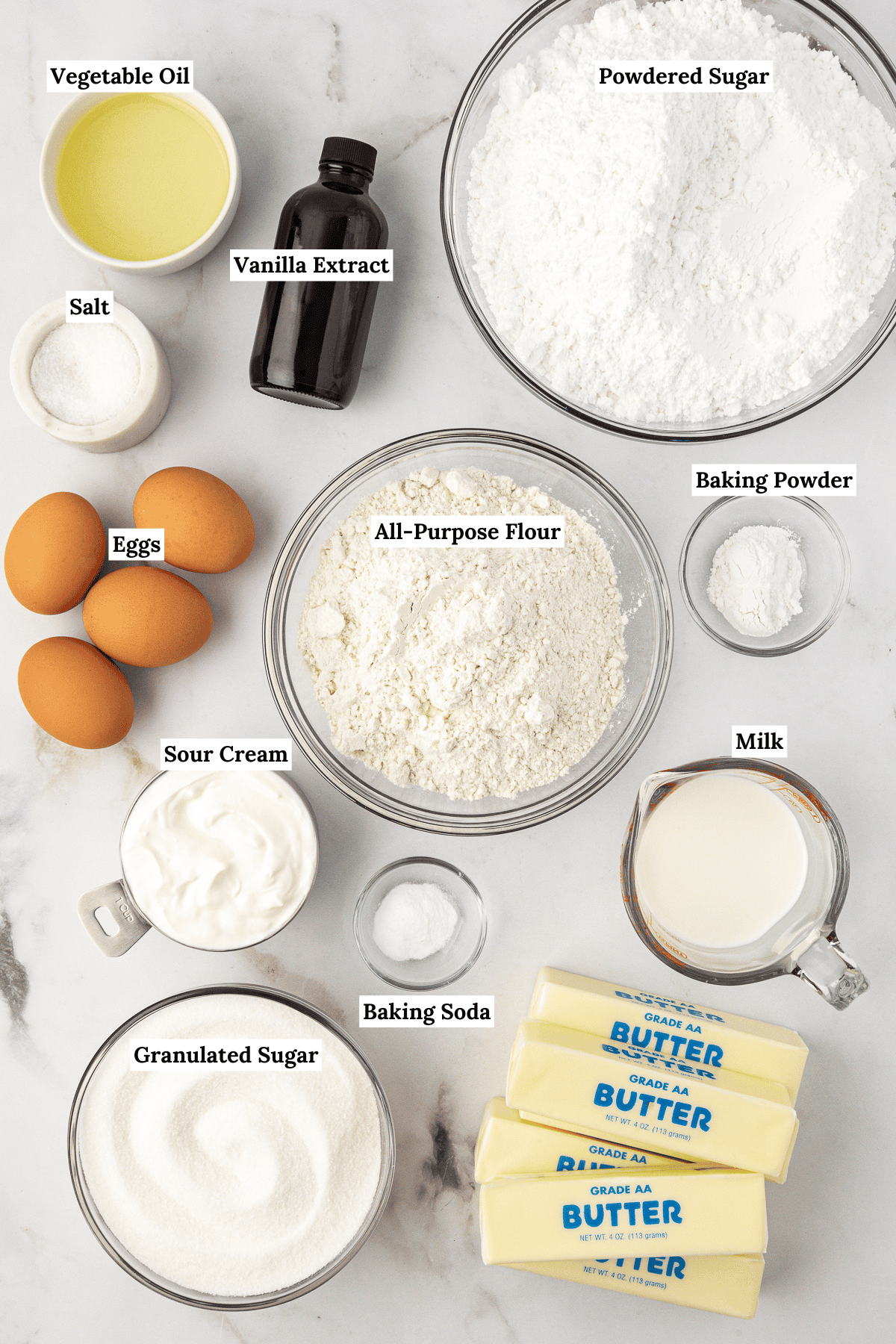 over head view of ingredients for vanilla cake including vegetable oil, powdered sugar, vanilla extract, salt, eggs, all-purpose flour, baking powder, sour cream, baking soda, milk, granulated sugar, and butter