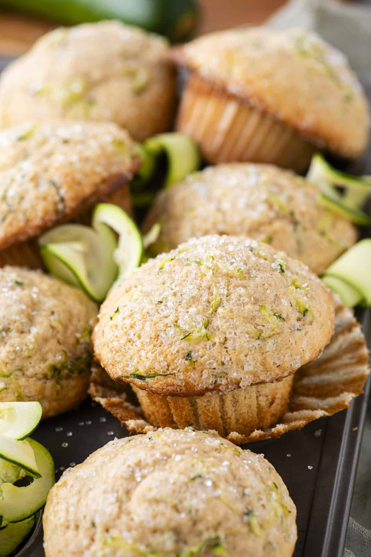 zucchini muffins piled on top of a muffin pan, the center muffin in the image sitting on top of it's peeled away muffin liner with rolls of thin zucchini slices between the muffins