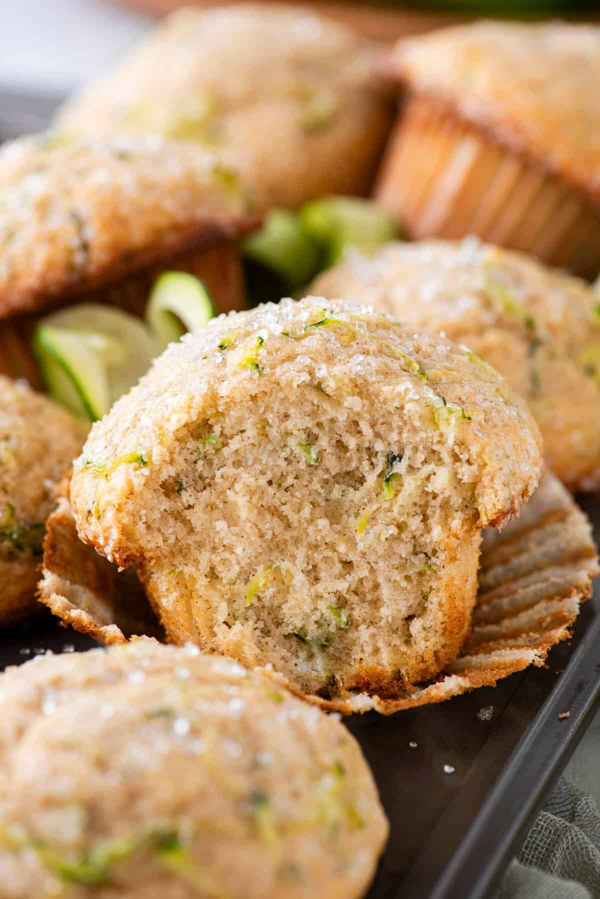 zucchini muffins piled on a muffin pan with the one in the center of the image peeled out of its muffin liner and a bite missing