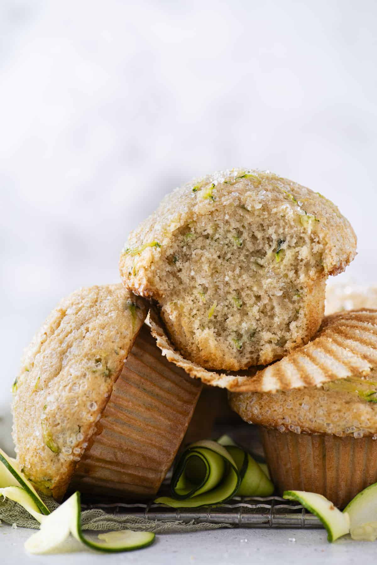 a pile of three zucchini muffins on top of a wire rack with thin zucchini slices scattered around, and the top muffin with its liner peeled away and a bite missing from the muffin