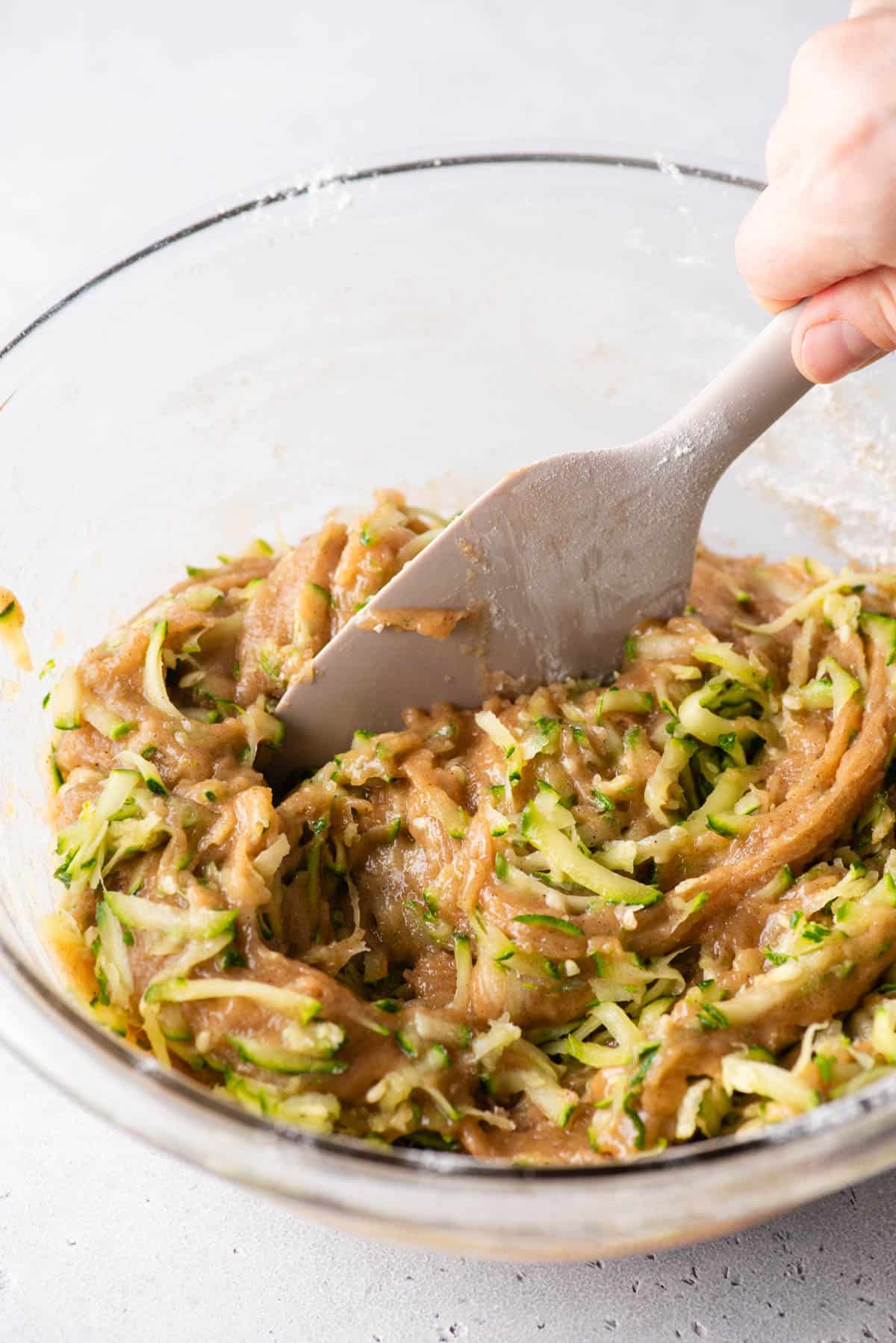a clear glass bowl with zucchini muffin batter and grated zucchini being mixed together with a spatula