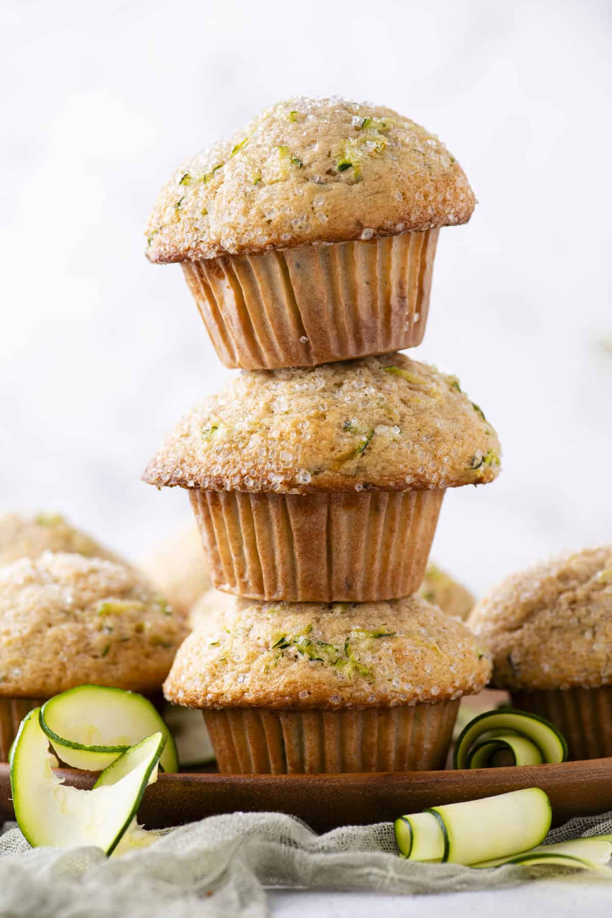 a tower of three zucchini muffins on top of each other on a wood plate with more muffins surrounding them and rolls of thin zucchini slices scattered around