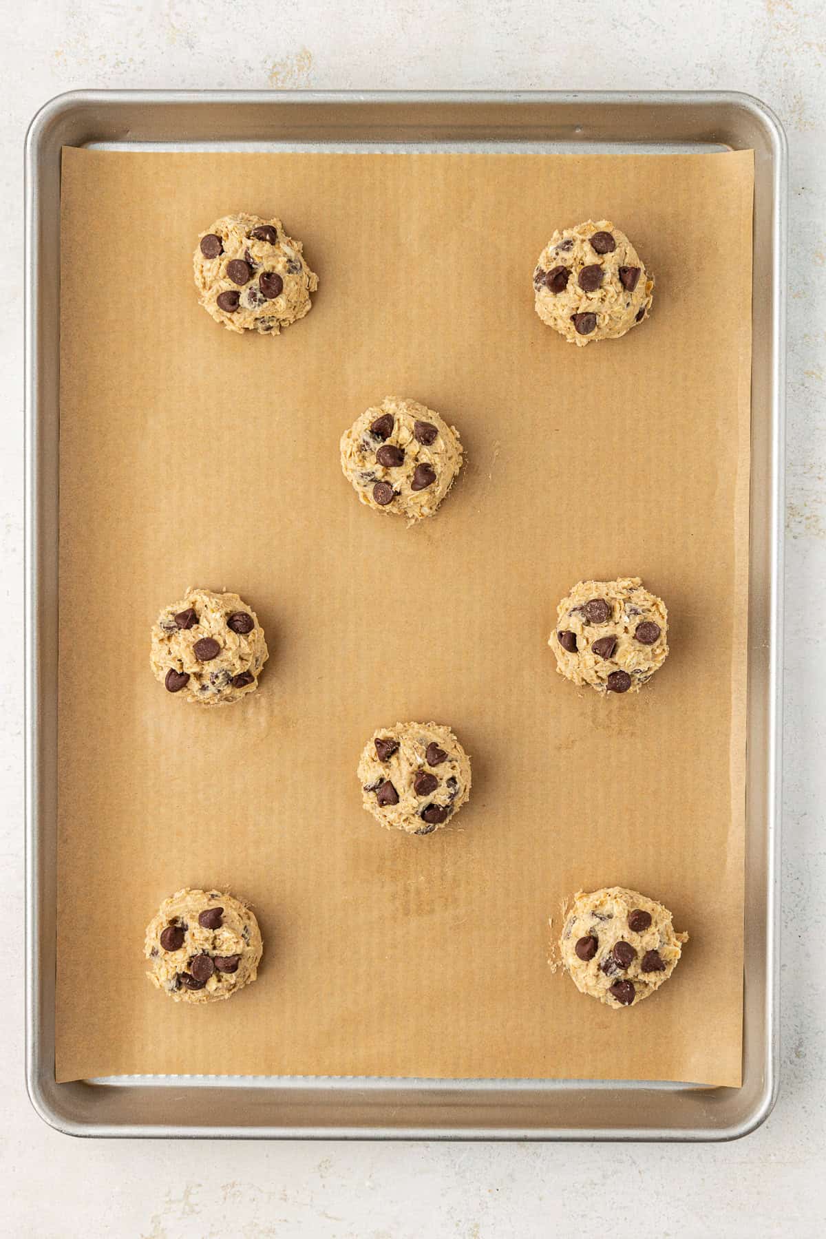 oatmeal chocolate chip cookie dough balls spread out on a baking sheet lined with brown parchment paper
